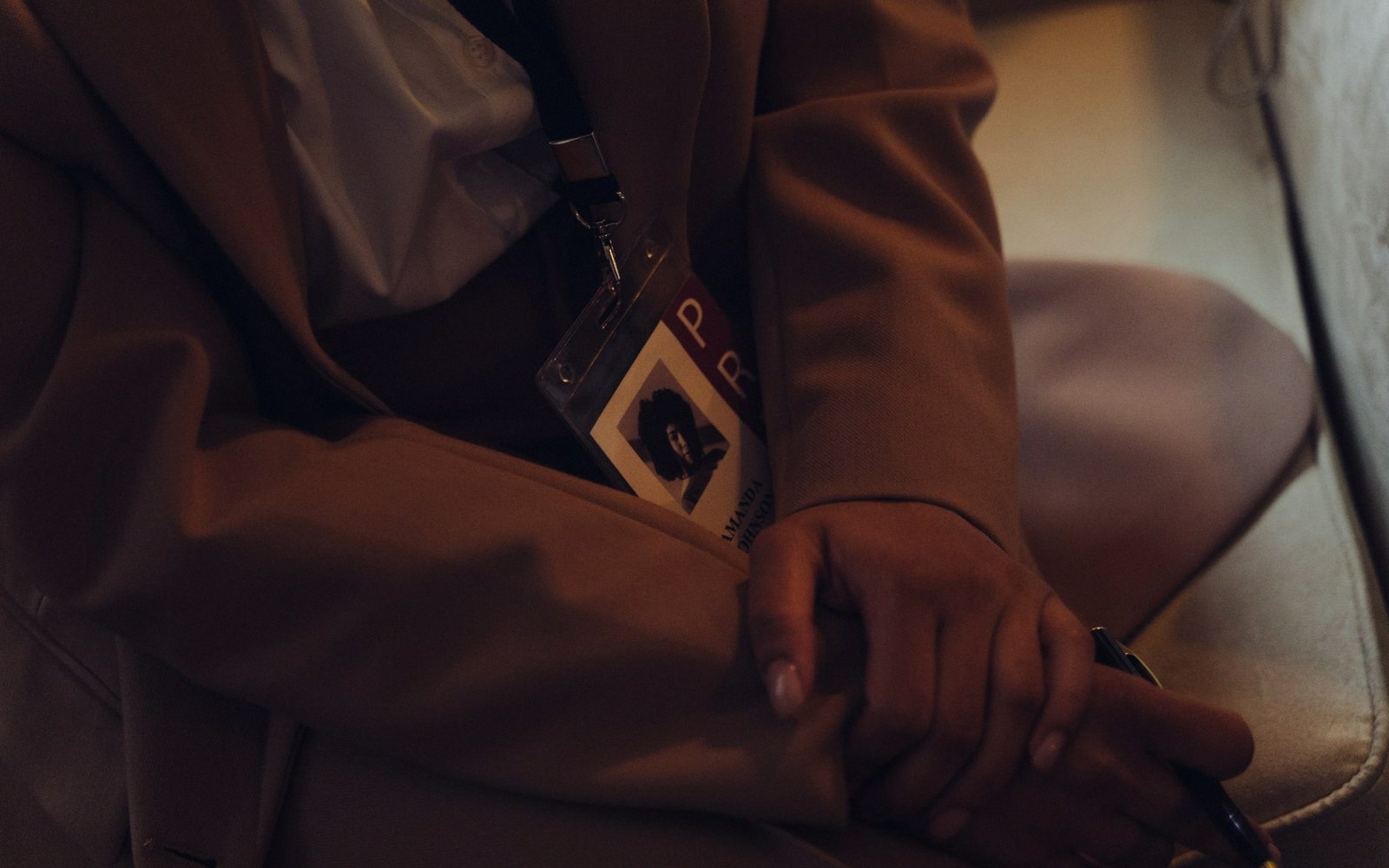 Seated person wearing a lanyard badge with clasped hands resting in their lap