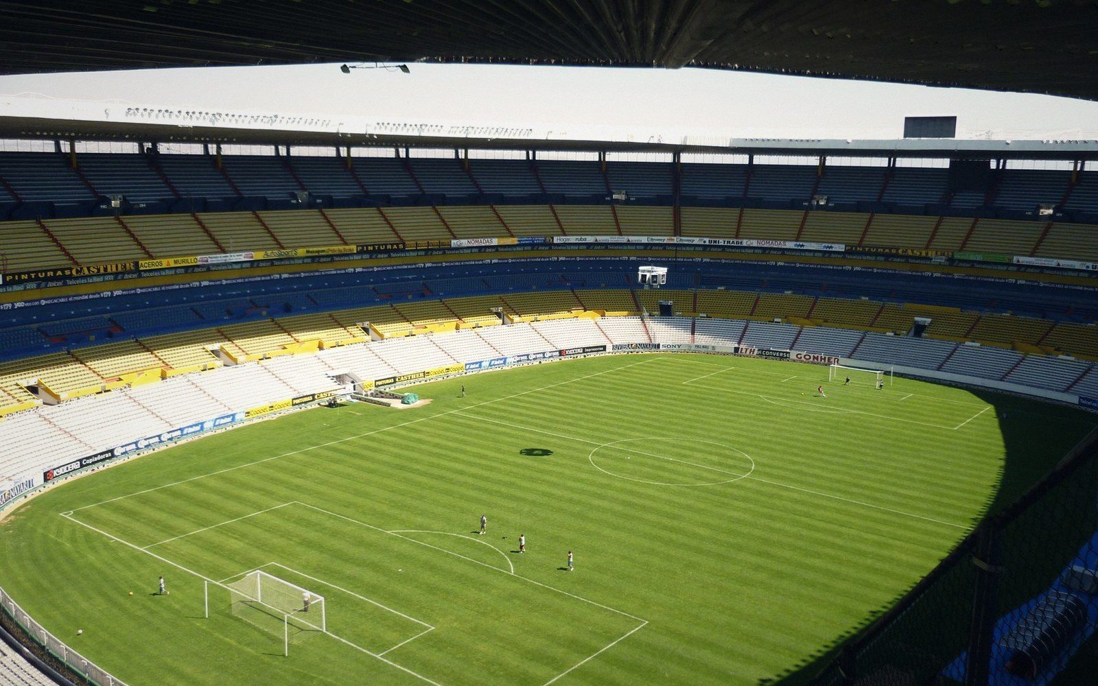 Empty stadium with tiered blue and yellow seating overlooks players training on the pitch