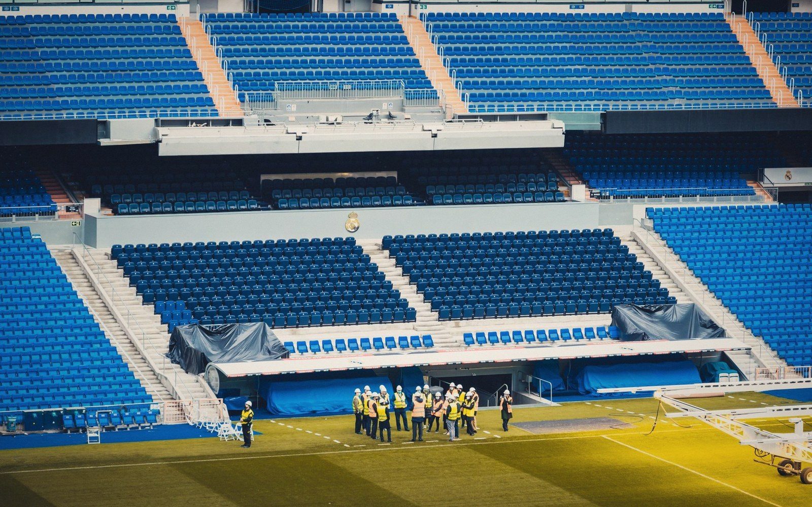 Workers in high-visibility vests gather on the pitch below empty blue stadium seating